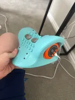 Imagem enviada pelo cliente. Clique para ver mais detalhes. A imagem representa: Um dispositivo eletrônico de massagem nos pés turquesa com furos e um display digital laranja sendo segurado na mão de alguém