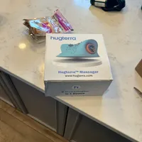 Imagem enviada pelo cliente. Clique para ver mais detalhes. A imagem representa: Uma caixa de produto Hugterra Massager colocada em uma bancada de mármore branco ao lado de alguns pacotes de salgadinhos e uma sacola preta