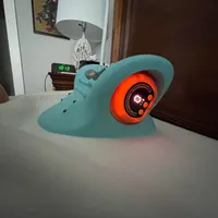 Imagem enviada pelo cliente. Clique para ver mais detalhes. A imagem representa: Um dispositivo eletrônico turquesa com uma tela circular laranja brilhante sobre uma mesa branca no interior de uma casa
