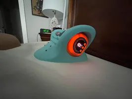 Imagem enviada pelo cliente. Clique para ver mais detalhes. A imagem representa: Um dispositivo eletrônico turquesa com uma tela circular laranja brilhante sobre uma mesa branca no interior de uma casa